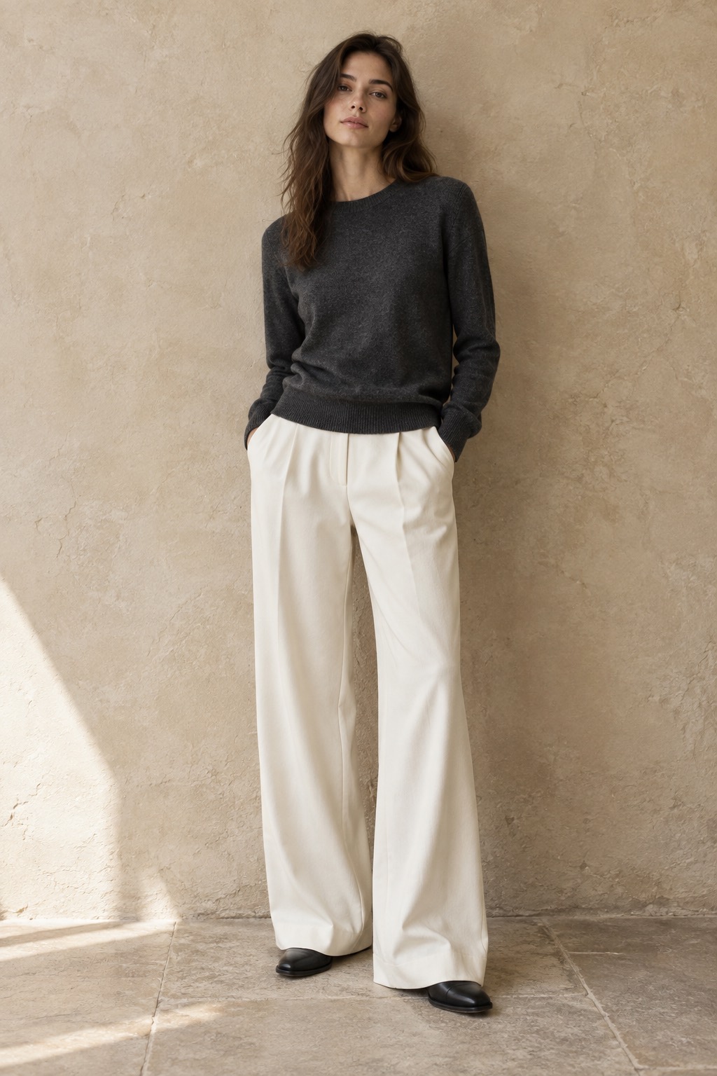A model in a charcoal fine-gauge knit sweater and ivory trousers against a plaster wall